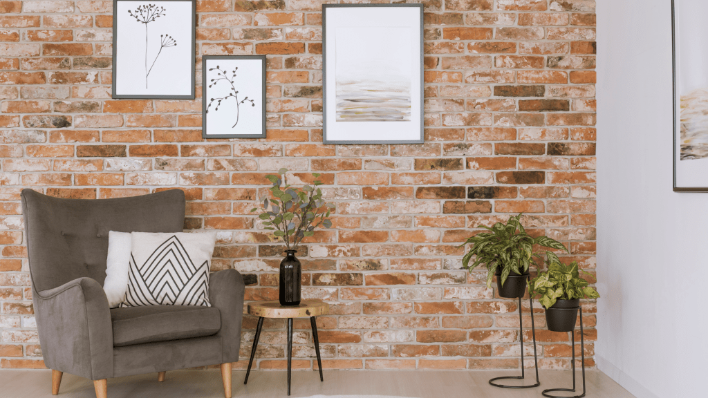 A living room set-up with 3 framed paintings on the brick wall, 3 plants in pots and a grey chair with 2 pillows.